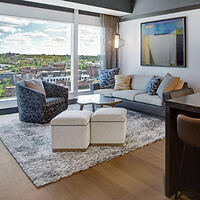 A modern living room with large windows offering a city view, a gray sofa, armchairs, and a plush rug.