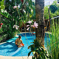 A person sits on the edge of a small, round pool in a lush, tropical garden with palm trees and pink flowers.