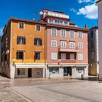 Three-story yellow and pink buildings with ground-floor shops face an empty paved square under a blue sky with scattered clouds.