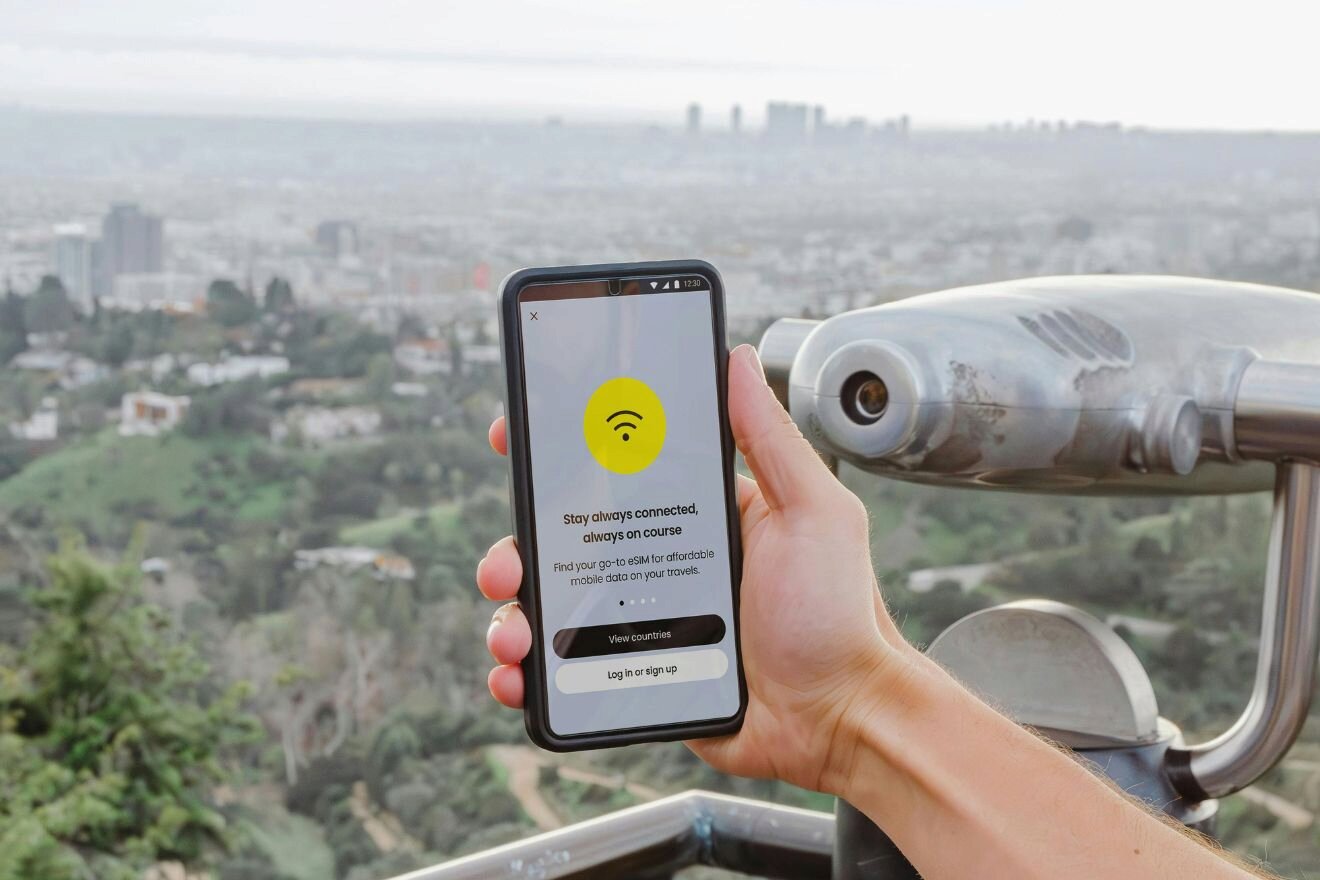A person holds a smartphone displaying a Wi-Fi connection app, with a cityscape visible in the background. The foreground includes part of a coin-operated binocular viewer.