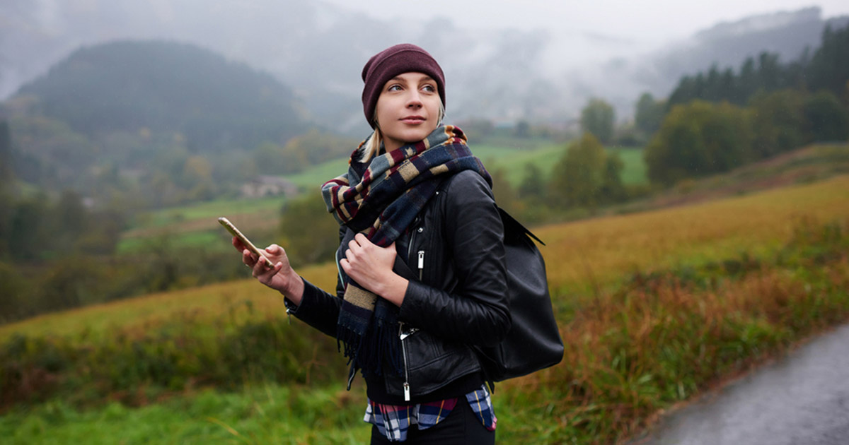 A person wearing a beanie, scarf, and leather jacket stands on a country road holding a smartphone, with a backpack on their shoulder. The background features misty hills and green fields.