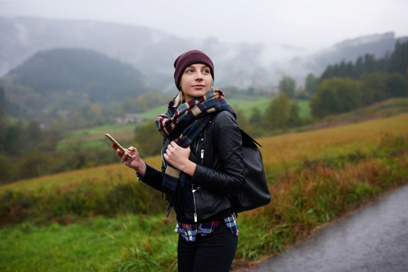 A person wearing a hat, scarf, and leather jacket holds a phone and stands on a path in a misty, mountainous countryside.