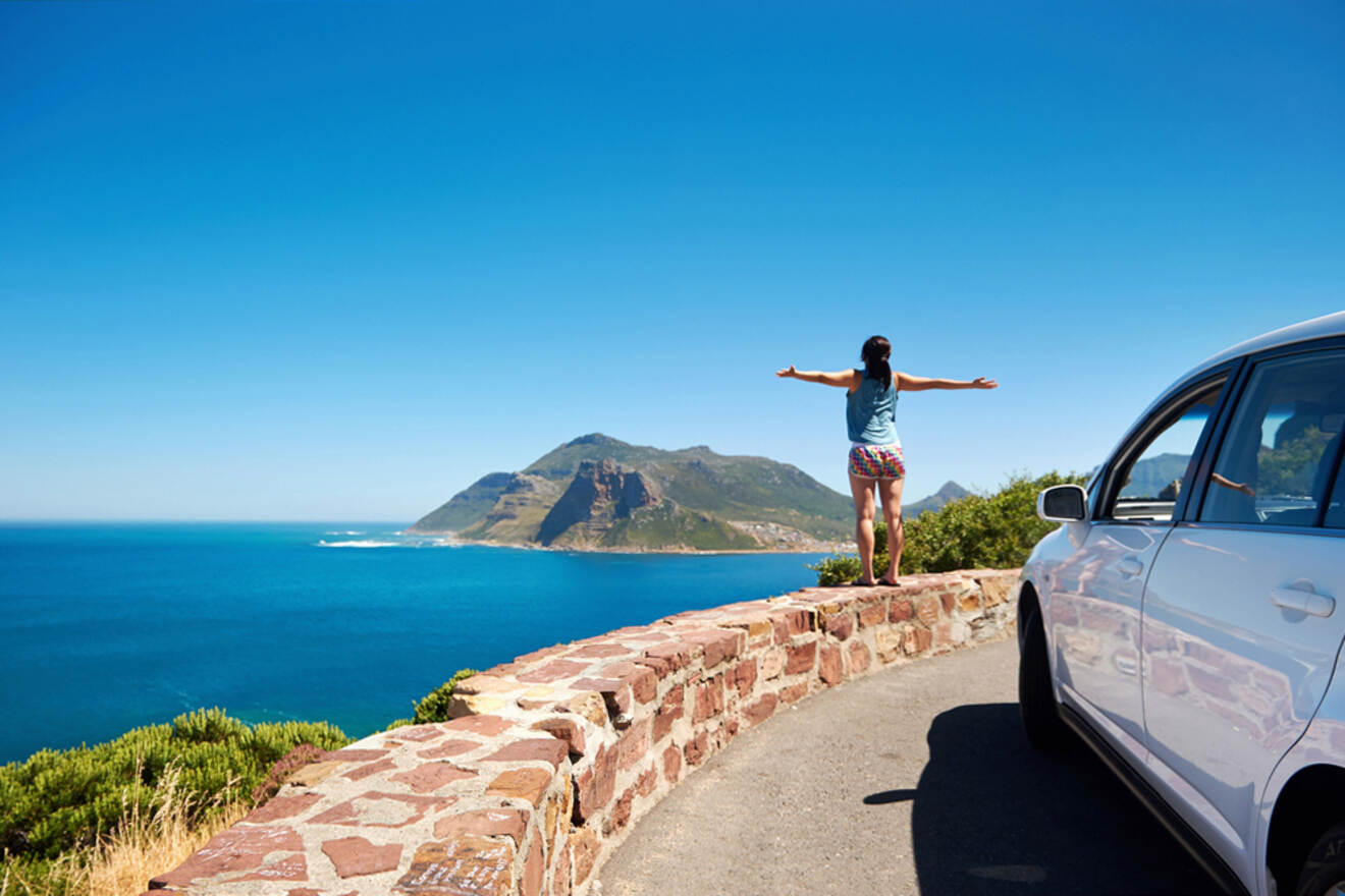 A person stands with arms outstretched on a stone wall, overlooking a coastal landscape with mountains and blue ocean, next to a parked white car under a clear sky.