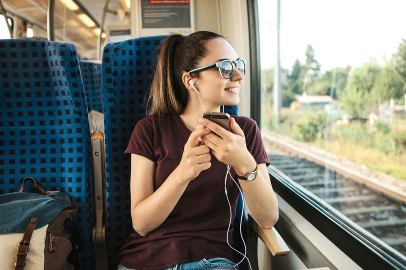 A woman with sunglasses and earphones is sitting on a train, holding a phone and looking out the window, smiling. A backpack is on the adjacent seat.