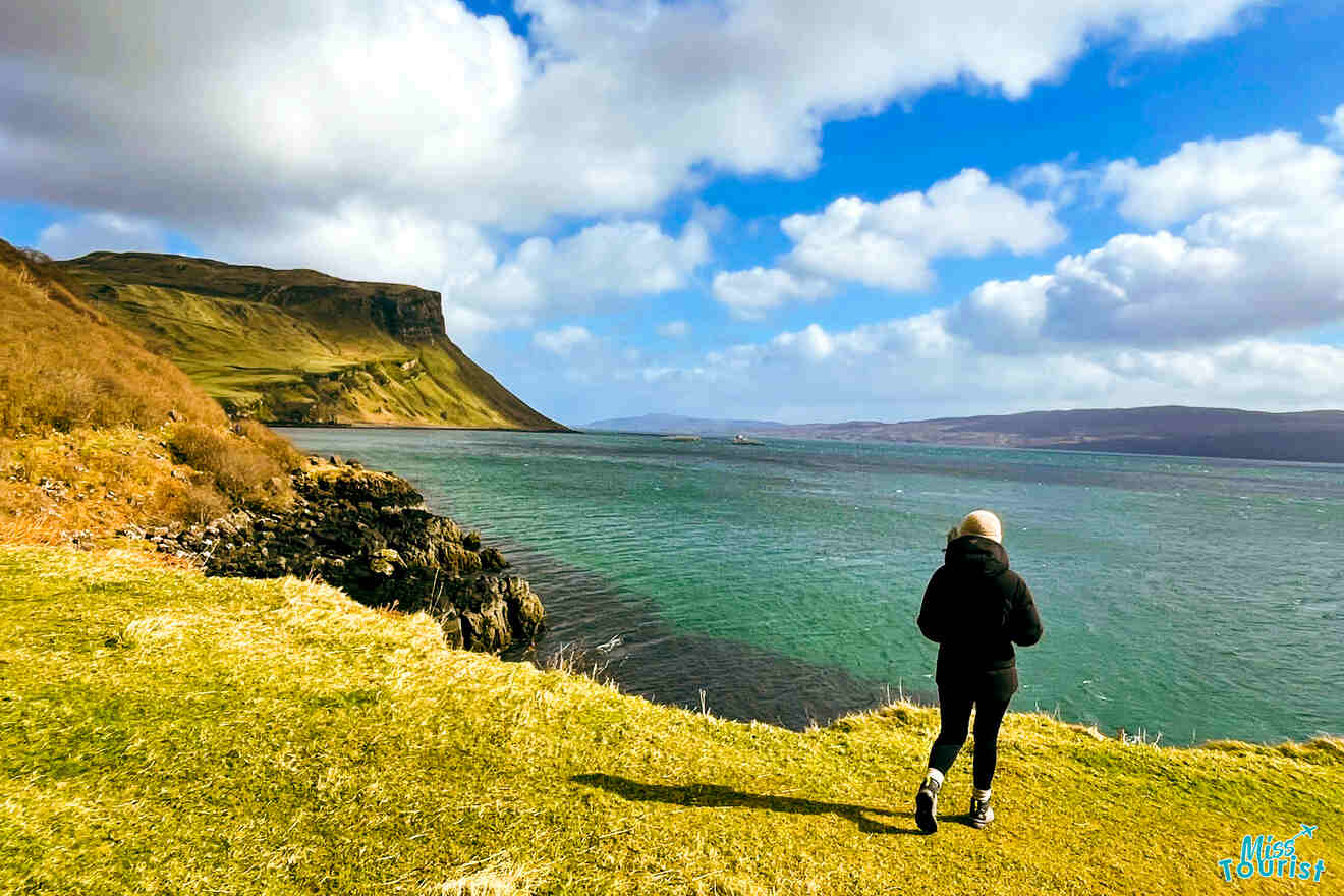Author of the post in a black jacket and hat walks near a grassy coastline overlooking a body of water with hills and cliffs in the distance under a partly cloudy sky.
