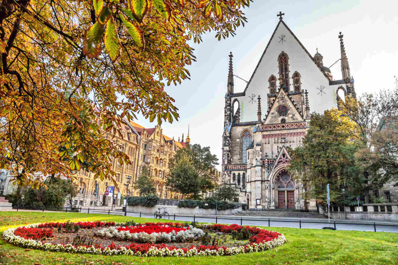 A historic church with a large, ornate entrance sits behind a circular garden with colorful flowers and vibrant autumn foliage.