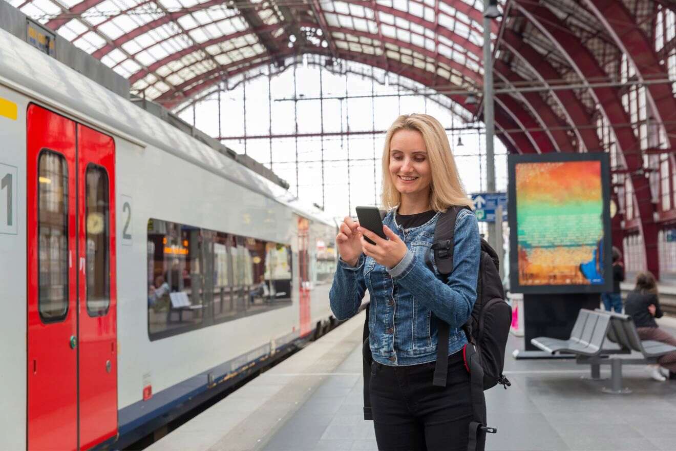 A person stands at a train station platform, looking at their smartphone and smiling. A train is stationary in the background.
