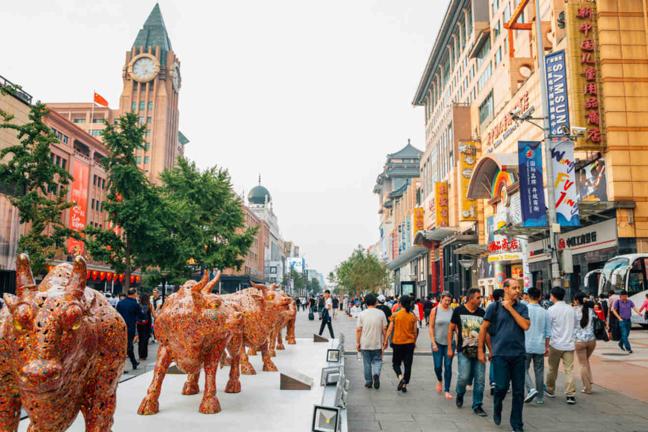 A bustling street in Beijing with people walking, colorful shops, and a line of artistic bull sculptures.