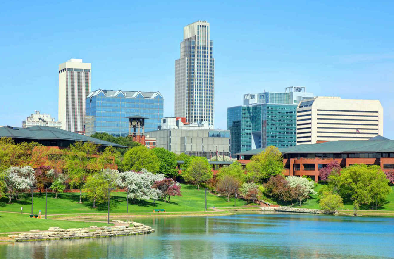 A scenic view of downtown Omaha with modern buildings, green trees, and a water body in the foreground