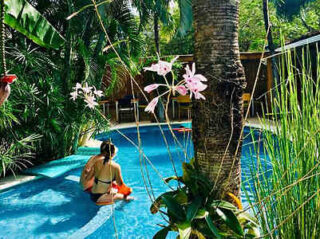 A person is sitting on the edge of a round swimming pool, surrounded by lush greenery and pink flowers, with a hut in the background.