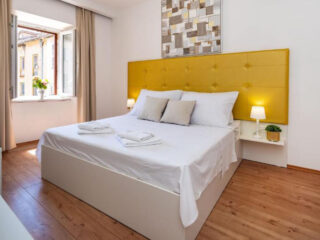 Um quarto moderno com cama de casal, cabeceira estofada amarela, lençóis brancos, mesas de cabeceira com abajures e uma janela aberta que permite a entrada de luz natural.