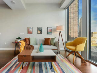 A bright living room with modern furniture, colorful decor, and large windows offering a city view.