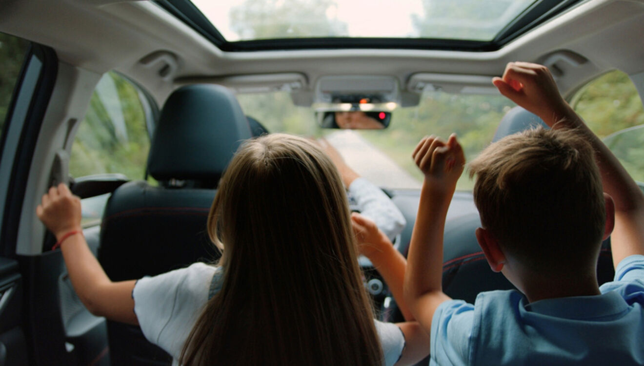Two children in the backseat of a car raise their hands in excitement, as the vehicle moves along a forested road.