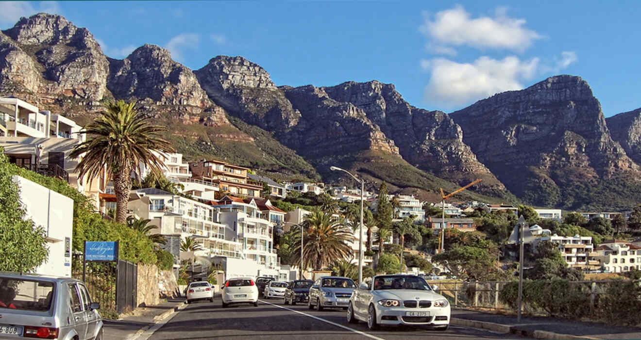 A hillside suburban area with numerous houses and palm trees, set against a backdrop of rugged mountain peaks. Several cars are driving and parked along the street.
