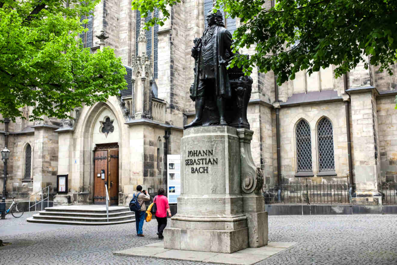 A statue of Johann Sebastian Bach stands in front of a historic church. Two people observe the statue, and trees frame the scene.
