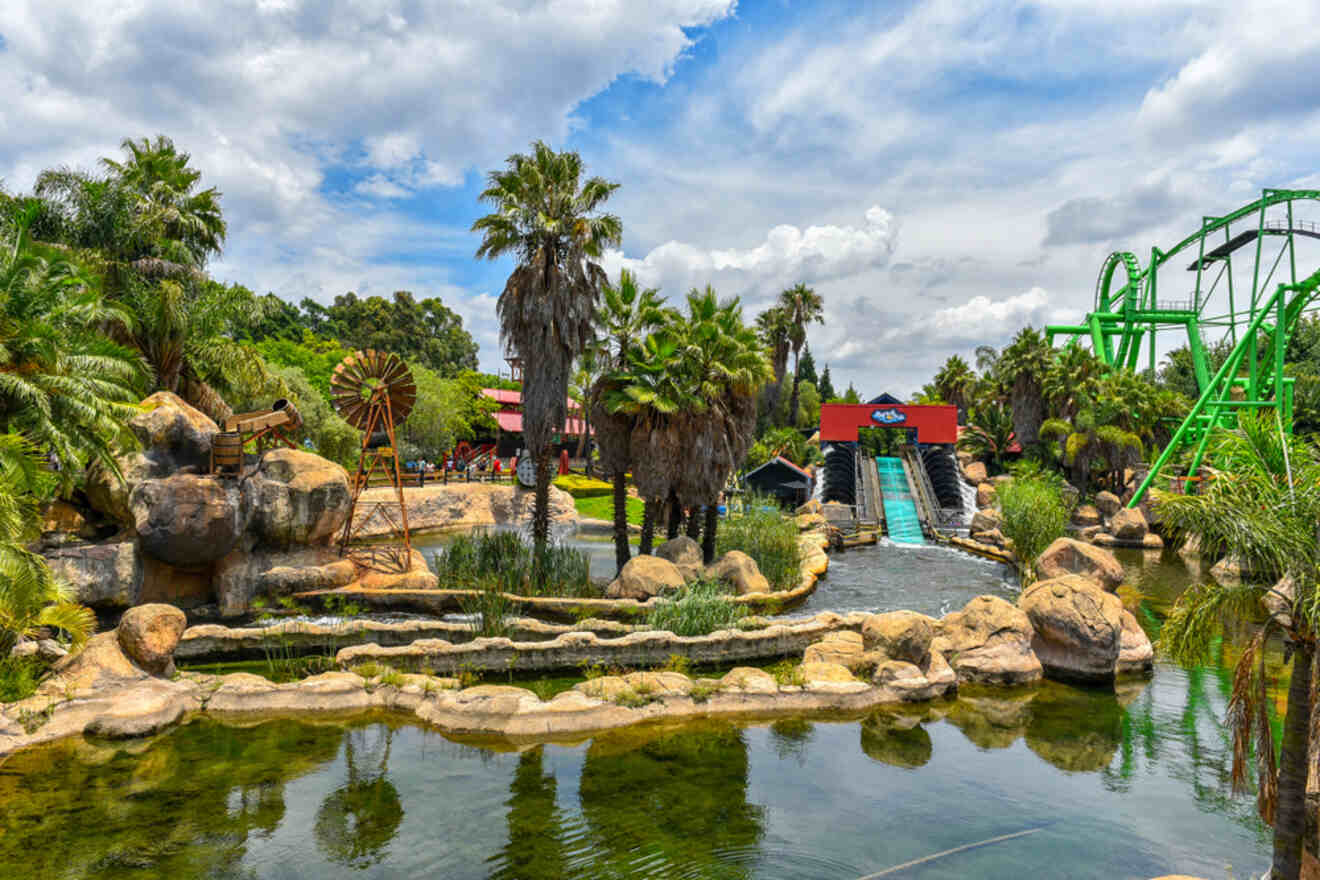 Amusement park with water rides, green roller coaster, and lush vegetation under a cloudy sky.