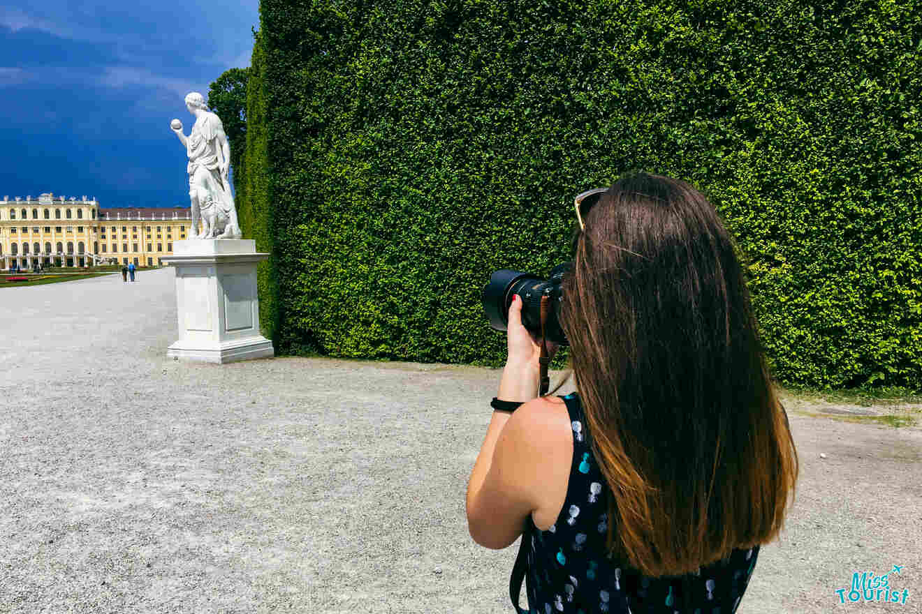 Author of the post with long brown hair is taking a photo of a white statue with a camera, in an outdoor setting with greenery and a grand building in the background.