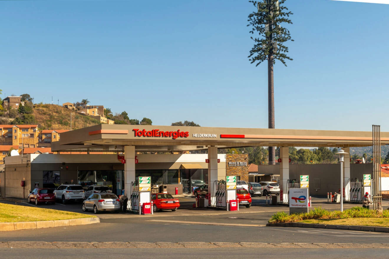 A TotalEnergies gas station with multiple vehicles fueling up. The station is located in an urban area with a single tall tree visible in the background.
