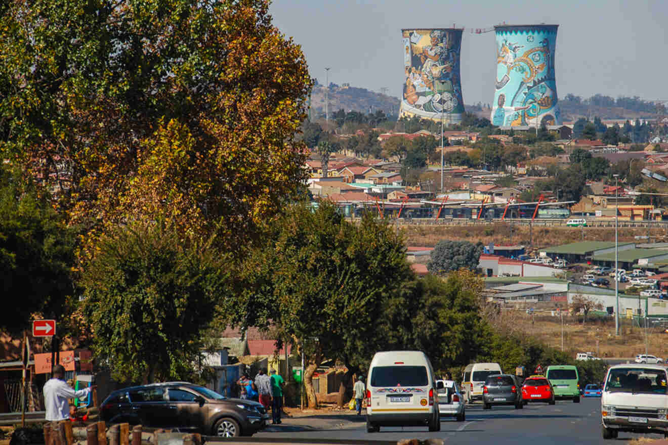 A street scene in Soweto with vehicles and pedestrians. In the background, two decorated cooling towers are visible, surrounded by residential buildings and trees.