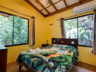 A small bedroom with yellow walls, a double bed with a tropical-themed bedspread, two large windows, and an air conditioner mounted above one window.