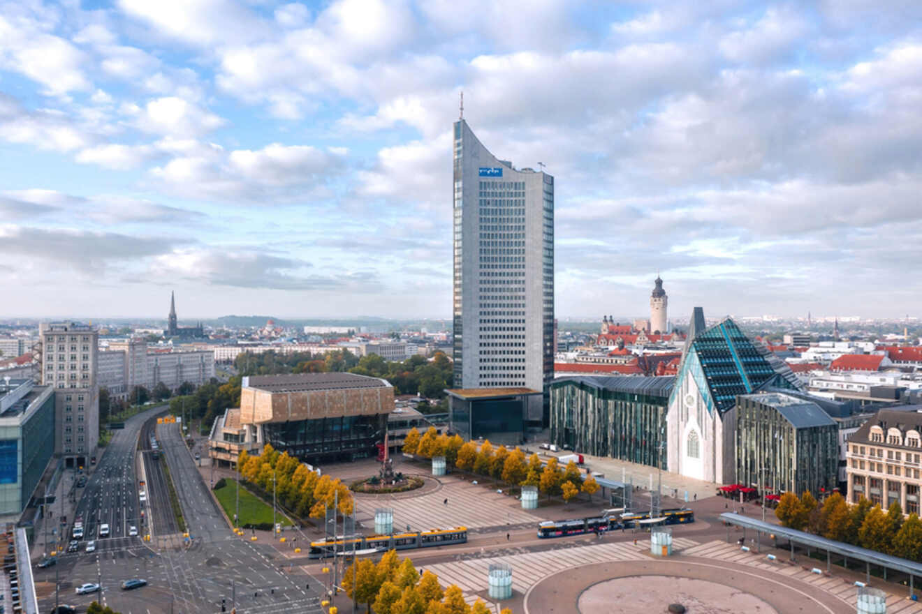 A cityscape of Leipzig, Germany, featuring a tall modern skyscraper, various buildings, an open plaza, and tree-lined streets. The sky is partly cloudy.