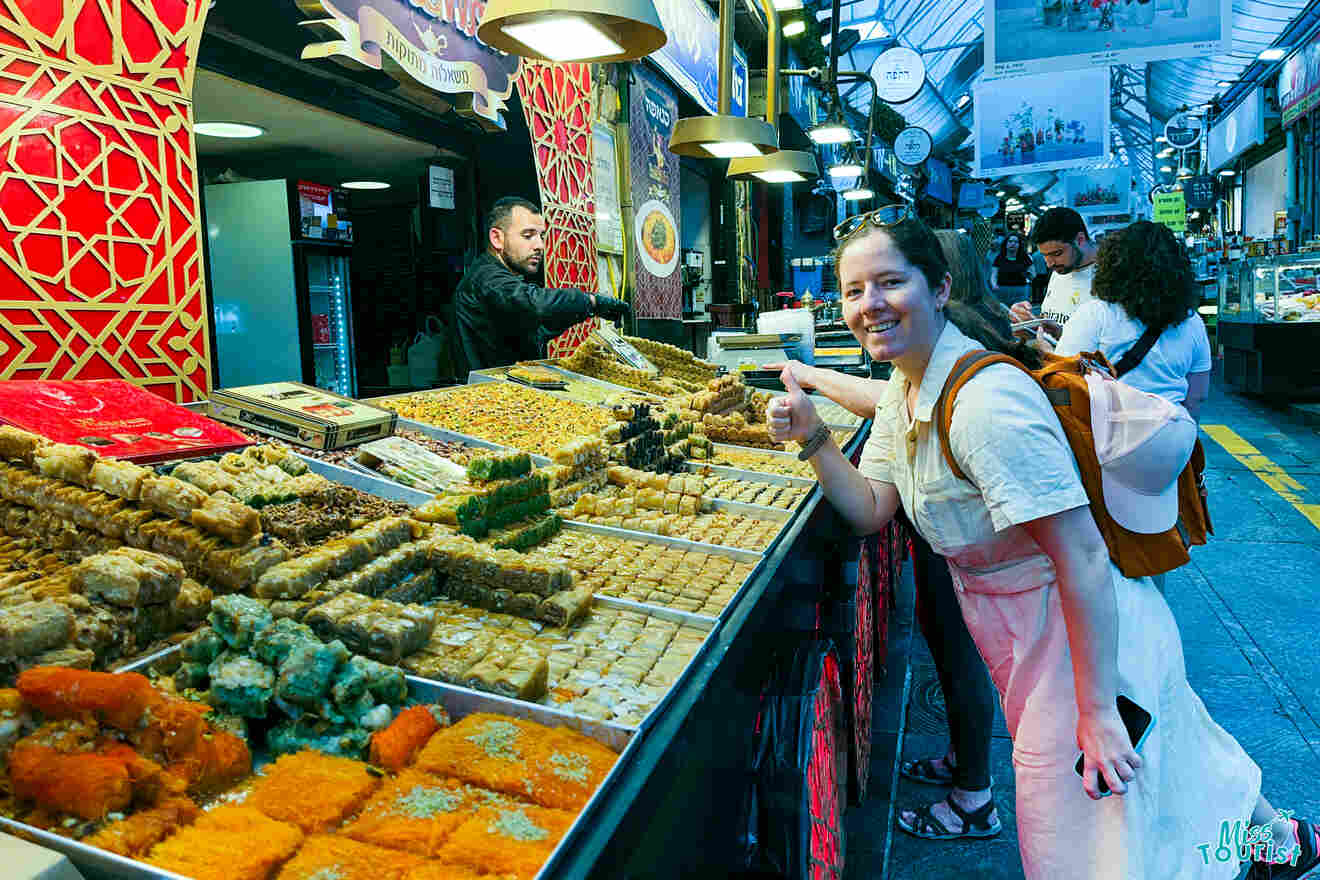 Author of the post with a backpack gives a thumbs up while standing next to a wide variety of pastries in a market stall. Other people are browsing the stall in an indoor market.
