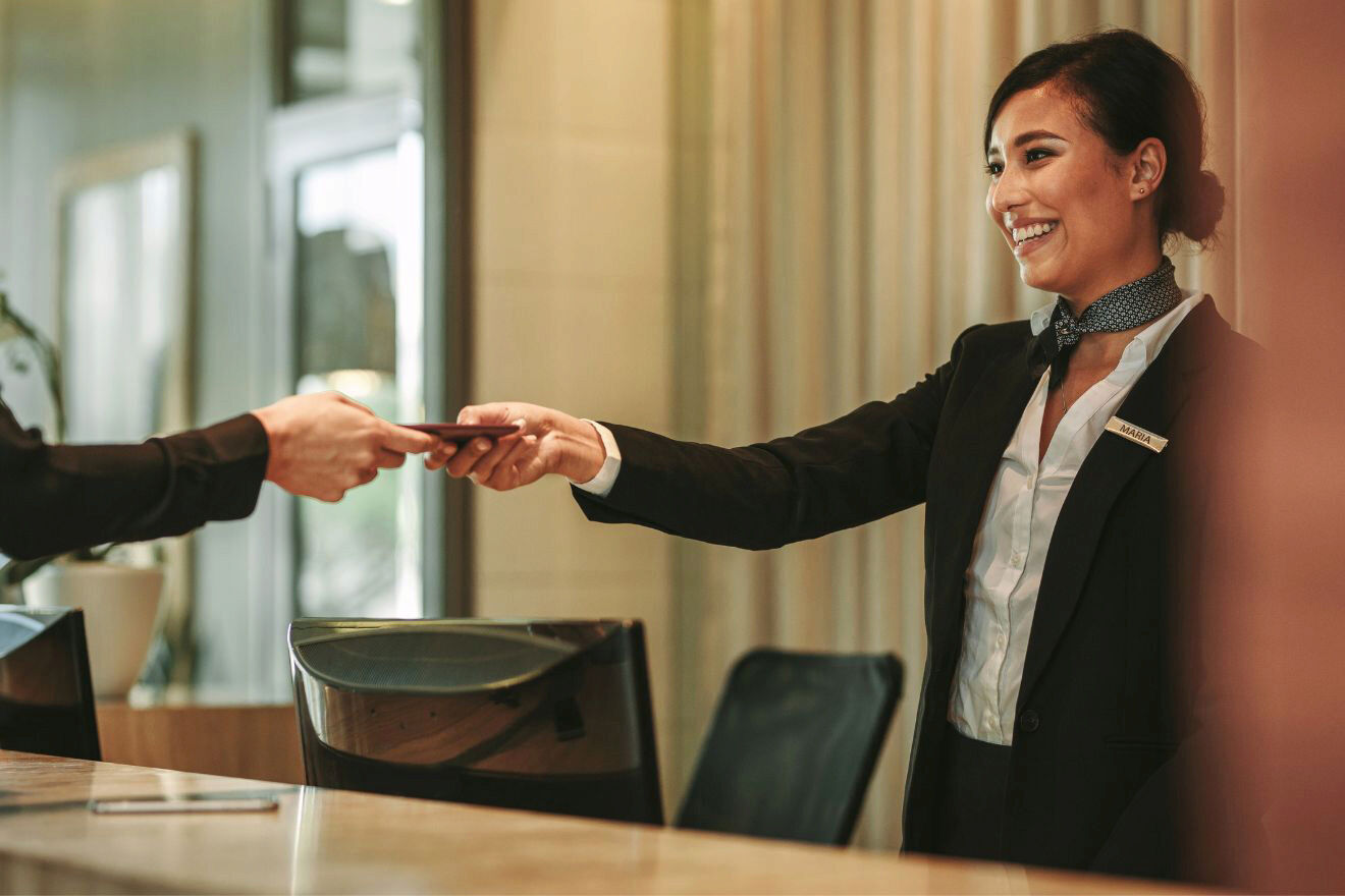 A hotel receptionist in a suit smiles while passing a document to a guest across the front desk.