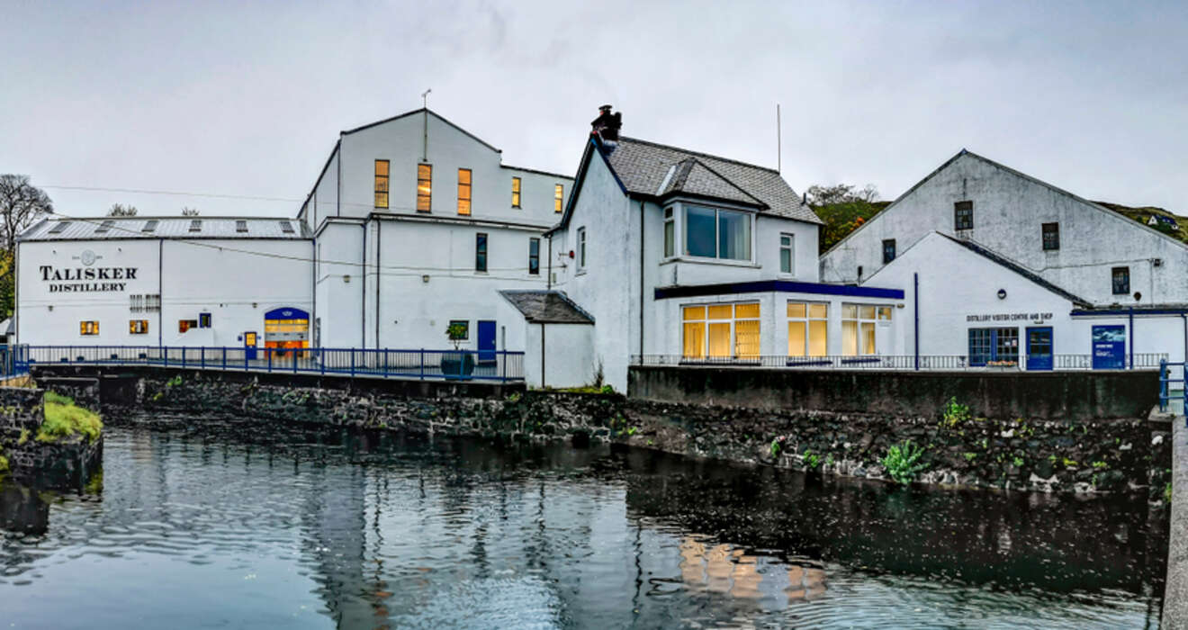 View of Talisker Distillery buildings next to a river, featuring white exteriors and lit windows, reflecting in the calm water. Overcast sky.