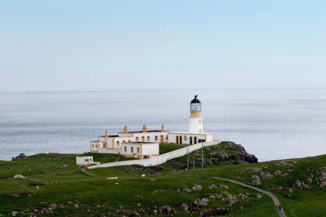 A white lighthouse with attached buildings sits on a grassy cliff overlooking a calm sea under a clear sky.