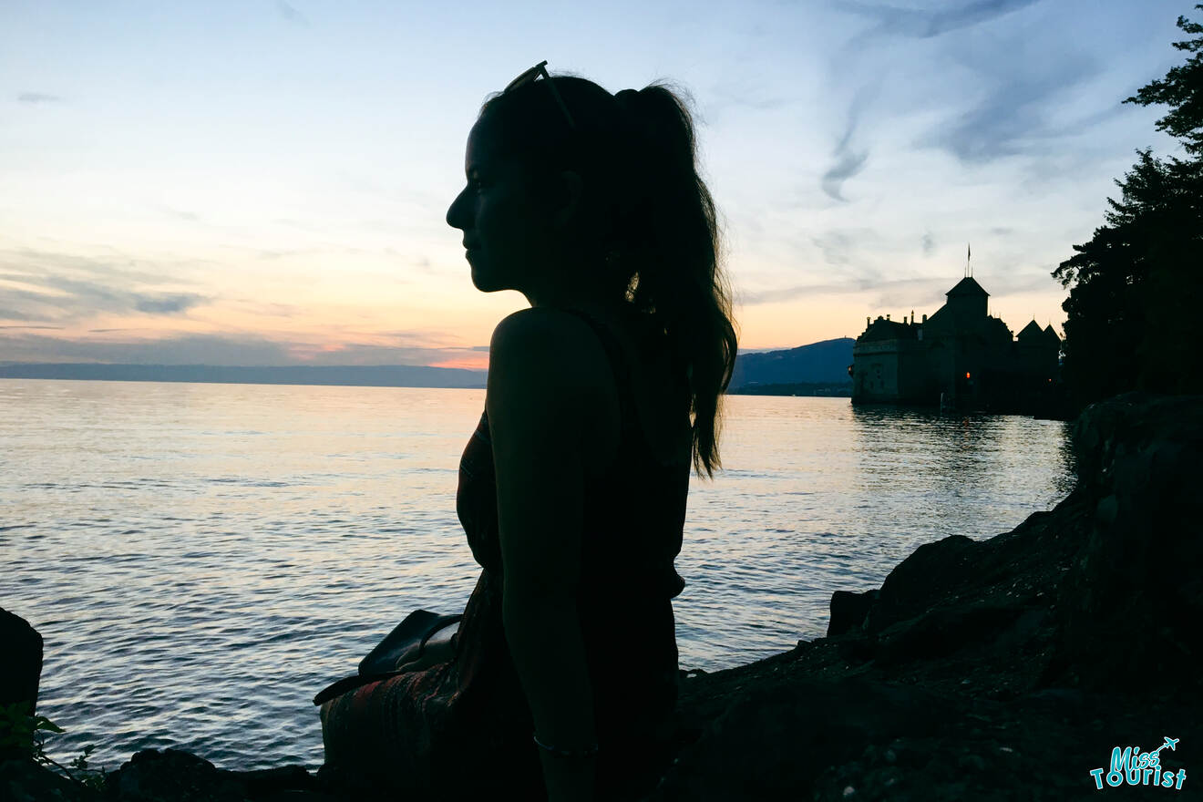 Silhouette of Author of the post sitting by the water's edge at sunset, with a distant castle-like structure visible in the background.