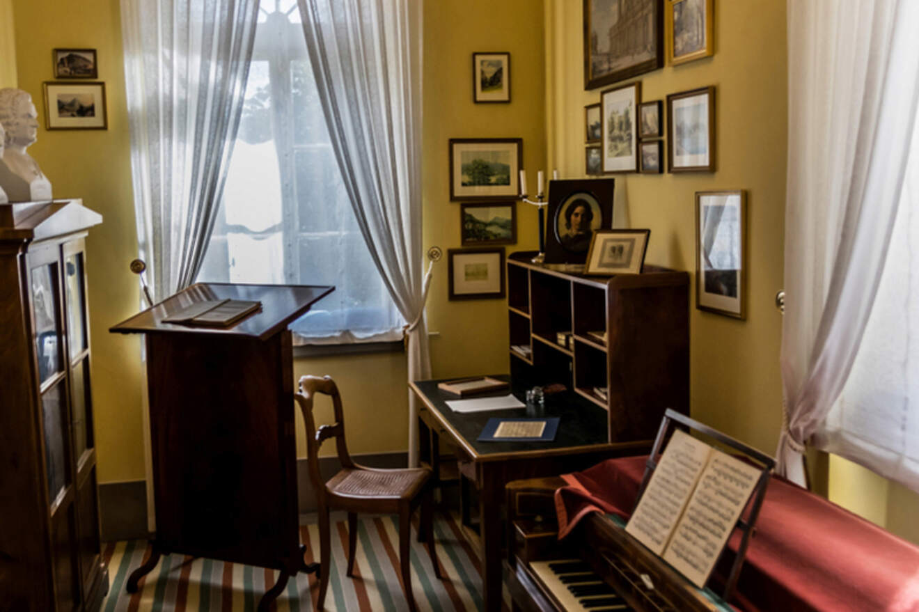 A vintage room setup features a writing desk, a chair, a piano with sheet music, framed pictures on the walls, and soft natural light coming through the window with lace curtains.