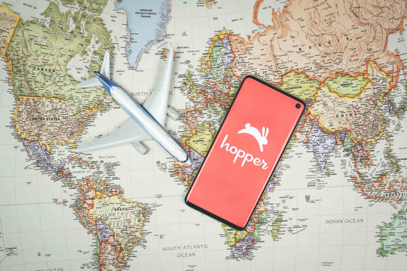 A smartphone displaying the Hopper app logo lies on a world map beside a model airplane.
