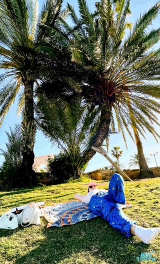 Author of the post lies on a blanket under two palm trees on a grassy area, wearing blue patterned pants and sunglasses. The sky is clear and sunny. There are bags and other items beside the blanket.