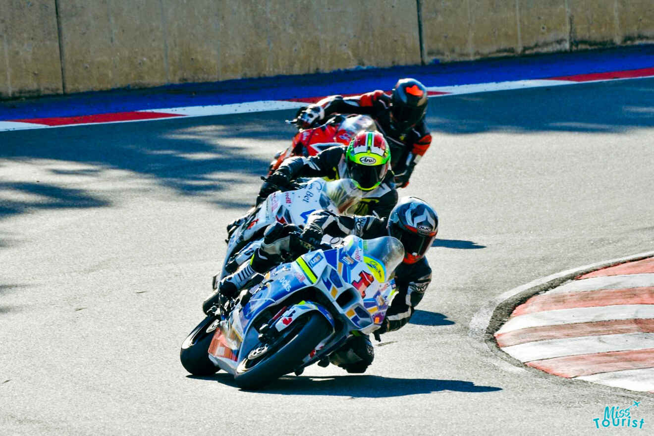 Four motorcyclists race closely around a bend on a track, leaning into the turn. The lead rider is followed closely by three others. The background shows a wall and racing lines on the track.