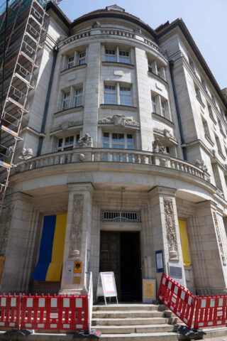 A multi-story stone building with a central entrance flanked by pillars. Scaffolding is on the left side, and a red barricade surrounds the entrance. Yellow and blue banners hang on either side of the door.