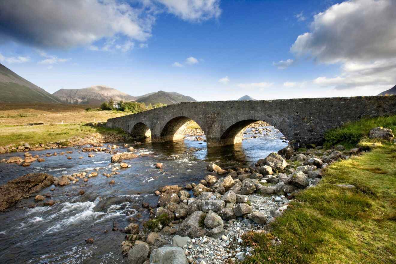 A stone bridge with three arches spans over a shallow, rock-strewn river, set against a backdrop of rolling hills and a partly cloudy sky.