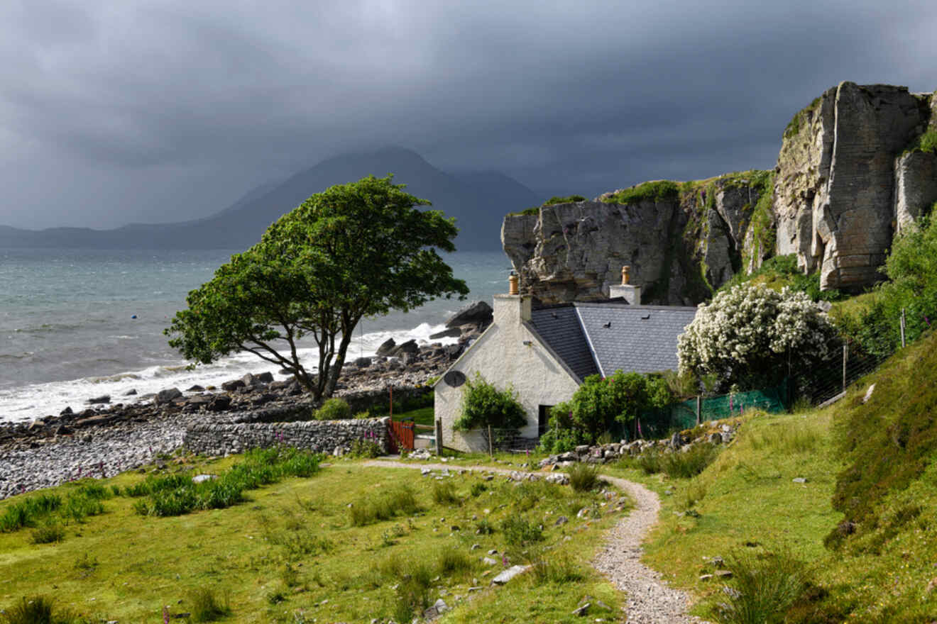 A stone house with a sloped roof is situated by a rocky shoreline, surrounded by greenery, a large tree, and cliffs; an overcast sky and distant mountains are visible in the background.