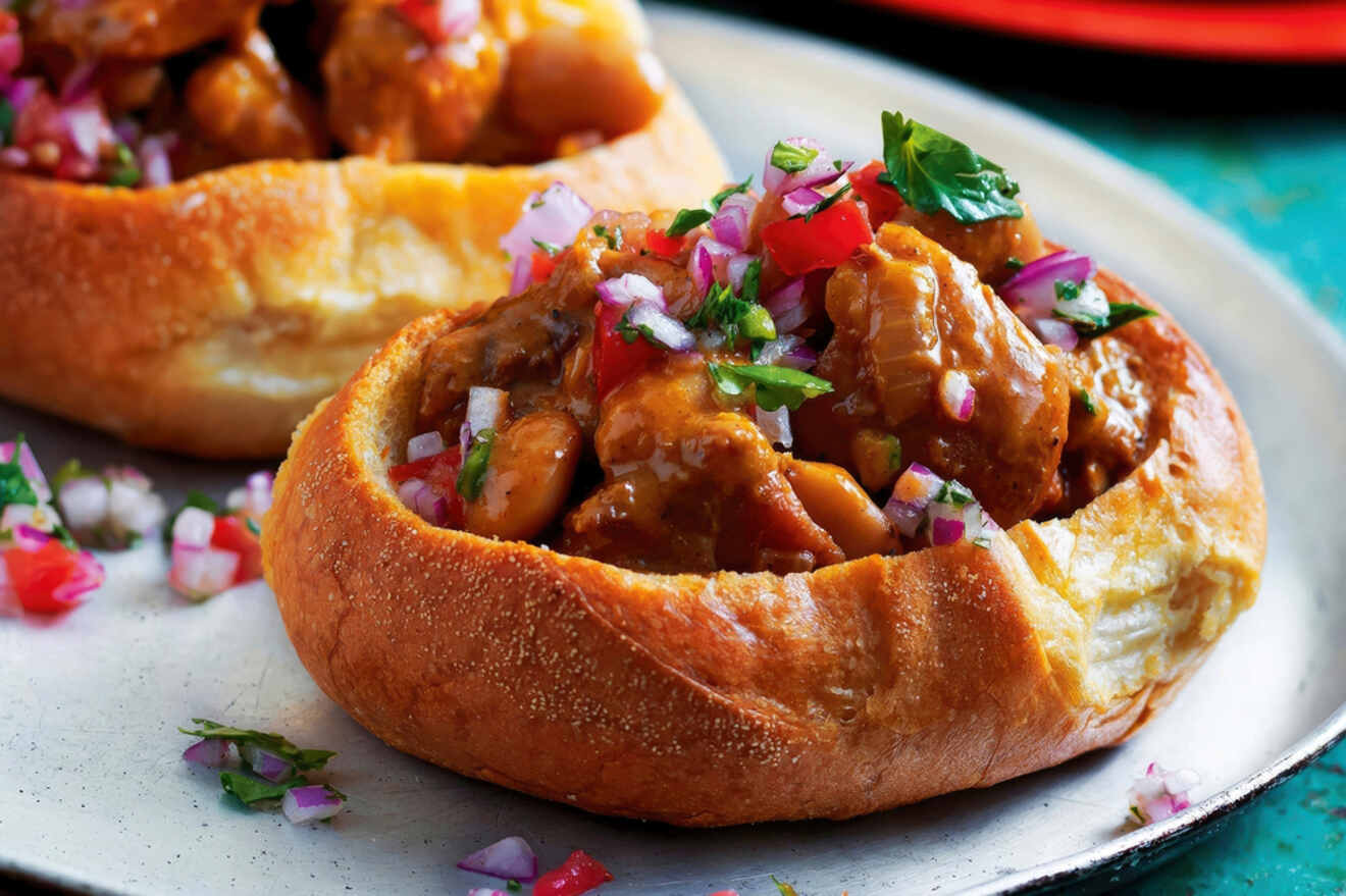 A bread bowl filled with a meat stew, topped with diced onions, tomatoes, and cilantro, is served on a plate.