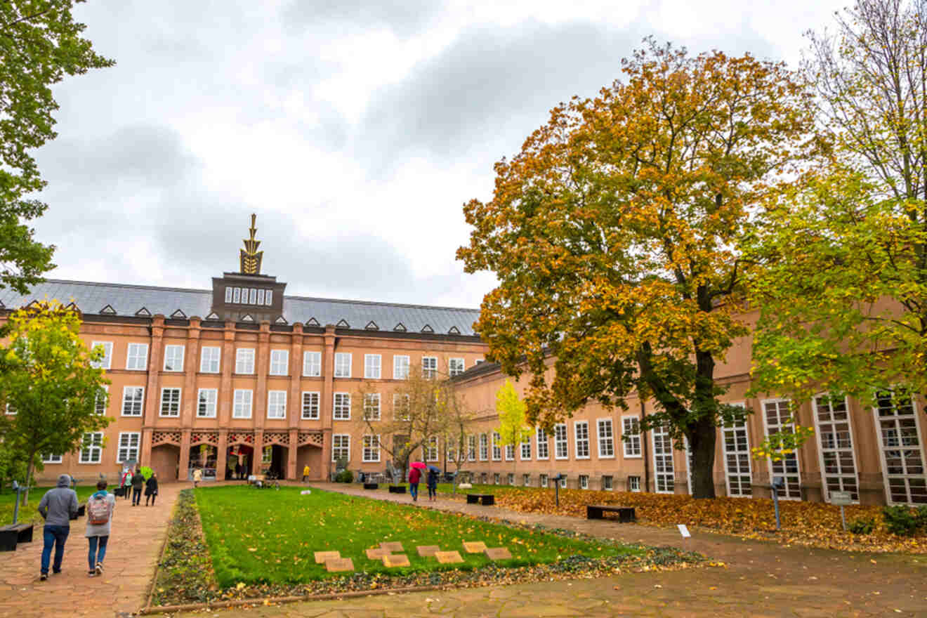 A group of people walking in the courtyard of a large brick building with autumn trees and overcast skies.