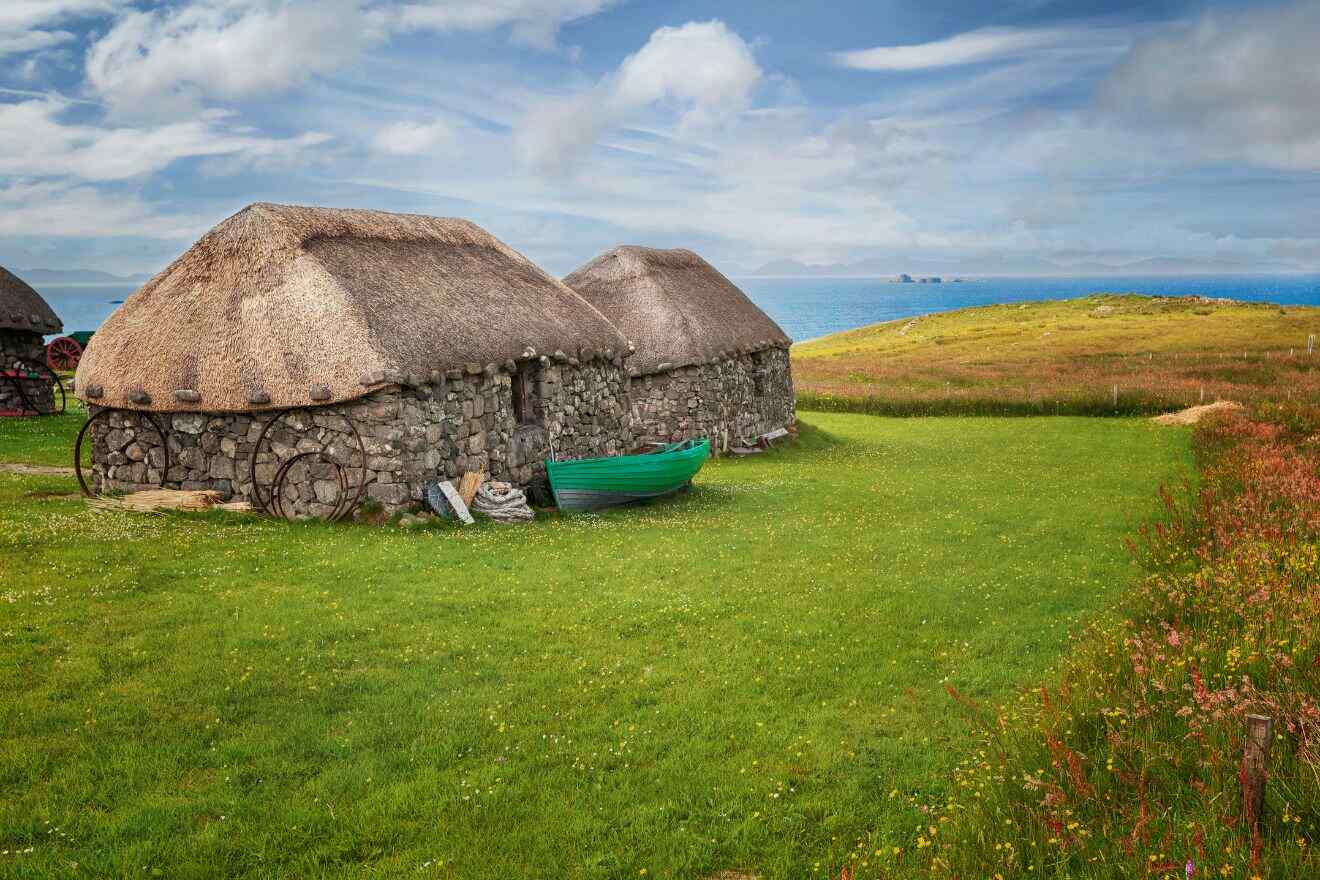 Two stone cottages with thatched roofs stand on a grassy field near the coastline, with a green boat and cartwheels placed beside them under a partially cloudy sky.