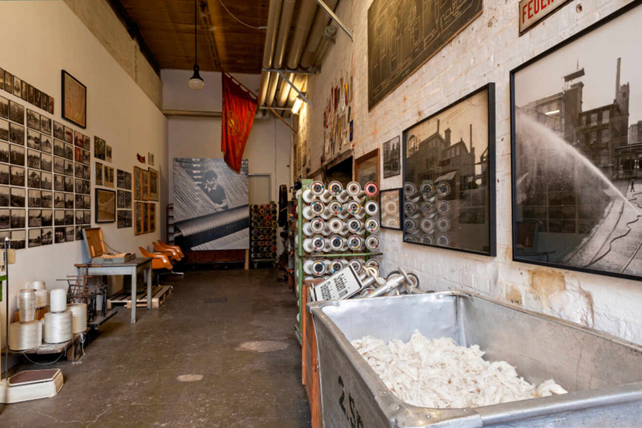 A hallway in a textile factory features yarn spools, historical photographs on the walls, and a cart filled with raw cotton. Industrial pipes and red safety signage are visible overhead.