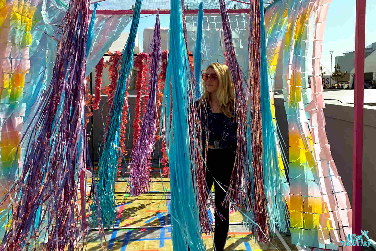 author of the post stands in a colorful installation with hanging ribbons in various shades, including blue and pink, while sunlight filters through.