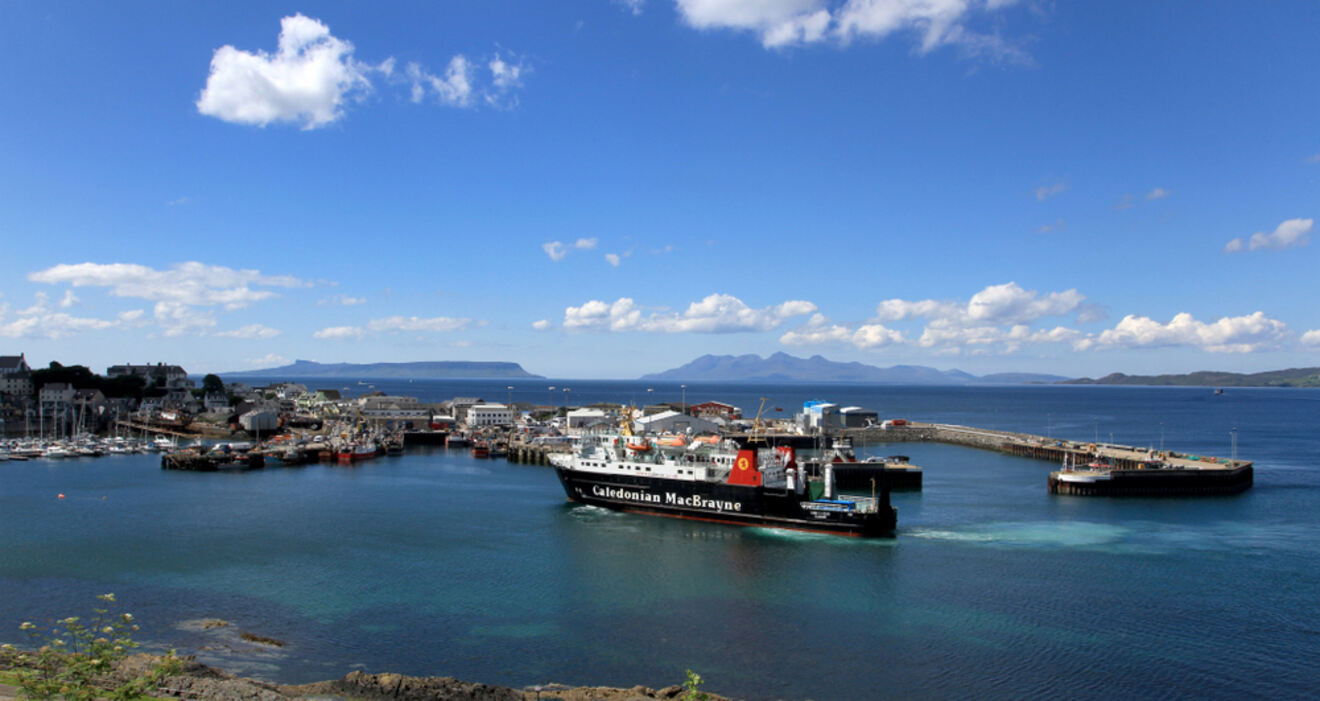 A ferry docked at a coastal harbor with other boats, surrounded by blue water and a clear sky with scattered clouds. Distant mountains are visible on the horizon.
