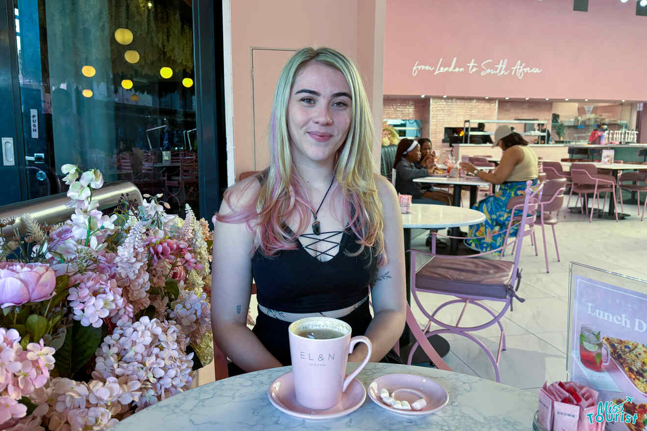 author of the post sits at a marble table in a café, with a coffee and small pastries in front of her. The café has a pink decor and other patrons can be seen in the background.