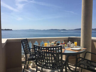 A dining table with a breakfast spread, including juice and pastries, on a balcony overlooking a calm ocean with distant hills and a passing boat under a clear blue sky.