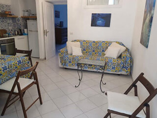A small living area with a patterned sofa, matching tablecloth, and simple wooden chairs, featuring a tiled floor.