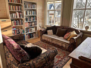 A cozy living room filled with bookshelves, patterned sofas, and large windows overlooking a snowy scene.