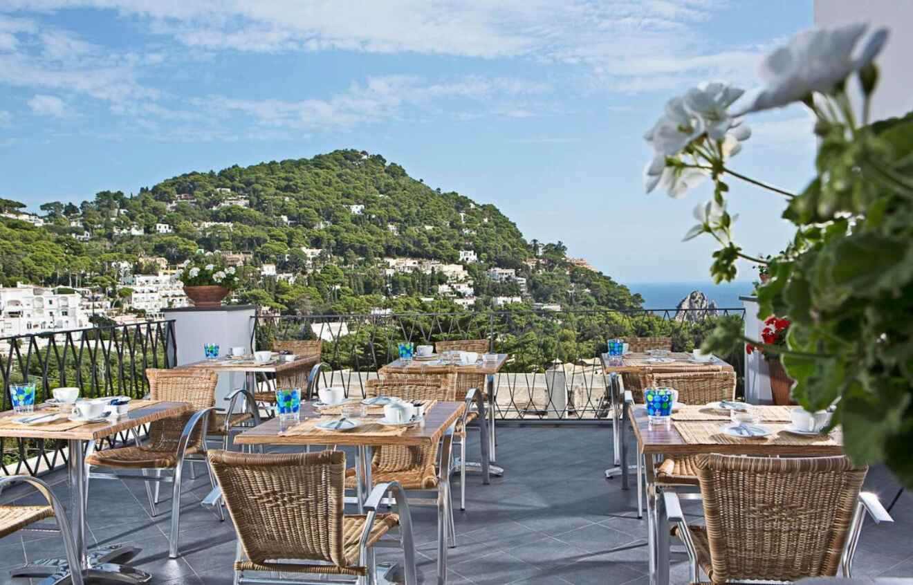 An outdoor terrace with wicker chairs and tables, offering a panoramic view of lush green hills and the sea in the distance