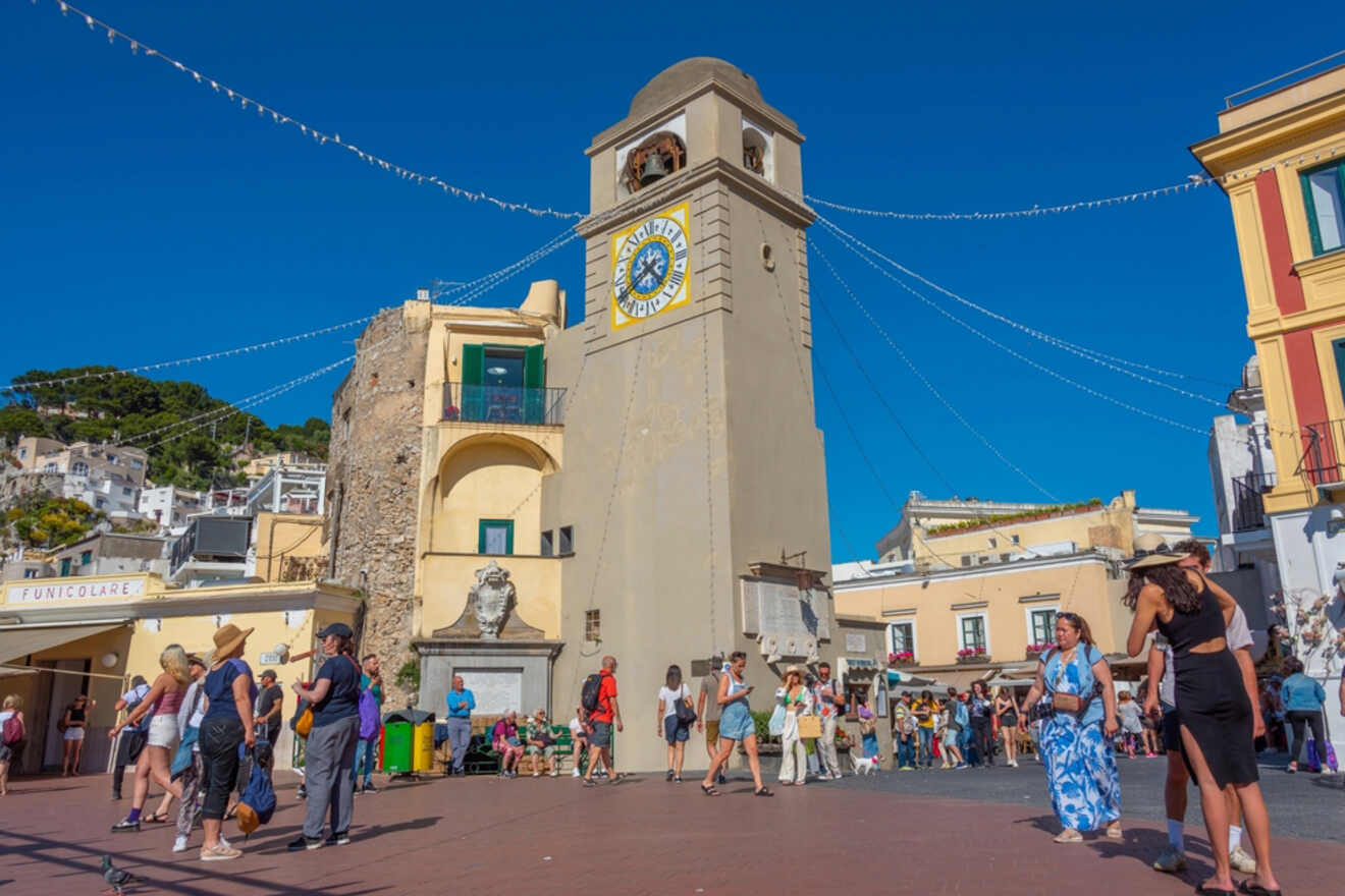 A bustling town square in Capri with people walking around, featuring a historic clock tower, colorful buildings, and a clear blue sky.