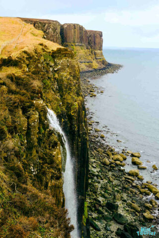Steep cliffs with a small waterfall cascading into the rocky shore below, meeting the ocean. Rugged landscape with sparse vegetation along the cliff tops. Overcast sky.
